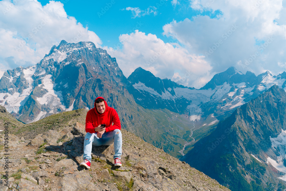 Man tourist sitting on rock with smartphone in mountains. Male traveler using mobile phone against cloudy sky on sunny day in mountainous terrain.