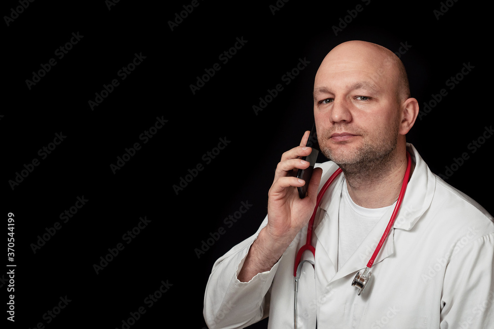 Portrait of a bald doctor in white uniform and red stethoscope on a black background. Model looking at the camera and holding phone. Portrait of a bald doctor in white uniform and red stethoscope on a black background. Model looking at the camera and holding phone.