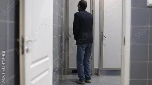 Man with urinary incontinence in public restroom near closed toilet cabin. Male person with frequent urination strongly wants to the restroom. Finally a womat going out from cabin
