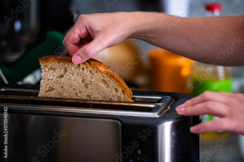 Female hand putting fresh whole wheat bread into toaster. Hands girl pulls in traditional slice bread. Blur background kitchen objects.