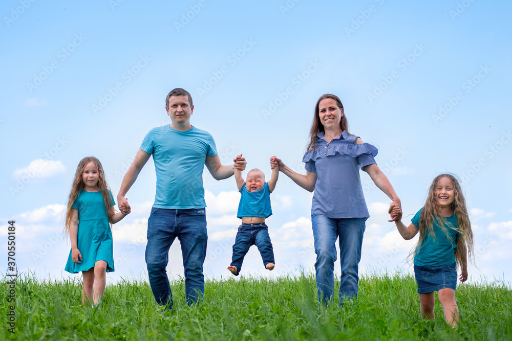 Big family dad, mom and three children walk on green grass against blue sky. Happy caucasian parents, two daughters and young son holding hands. Big family dad, mom and three children walk on green grass against blue sky. Happy caucasian parents, two daughters and young son holding hands.