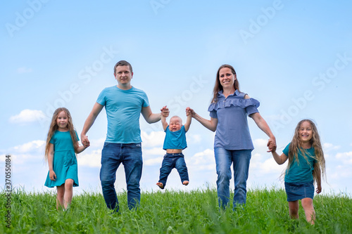 Big family dad, mom and three children walk on green grass against blue sky. Happy caucasian parents, two daughters and young son holding hands.