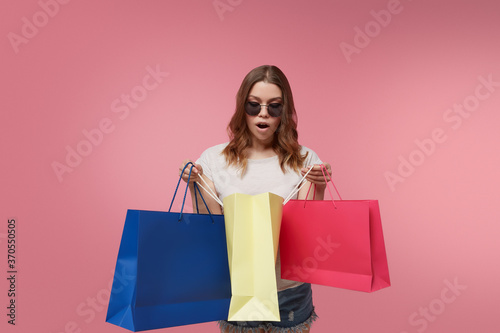 Shopping as a kind of a leisure activity and an effective stress reliever. Admired happy lady in sunglasses, denim shorts and white t-shirt is looking into colorful shopping bags in her hands.