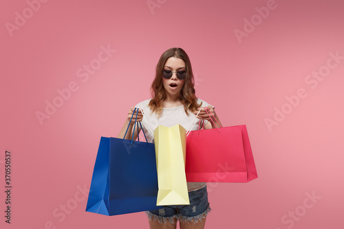 Shopping as a kind of a leisure activity and an effective stress reliever. Admired happy lady in sunglasses, denim shorts and white t-shirt is looking into colorful shopping bags in her hands.