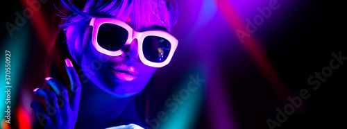 Disco girl in neon light dancing in night club. Fashion model woman. Closeup portrait of beautiful girl with fluorescent make-up. Body Art design in UV. Sunglasses. Colorful make up