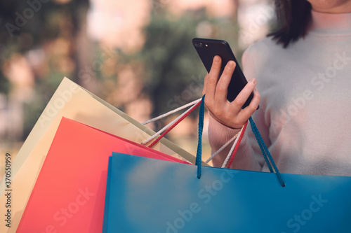Online shopping saves money, time and helps to relieve the stress. Cropped shot of a lady in white pullover and with a phone and colorful shopping bags in her hand. Outdoor photo on blurry backdrop.