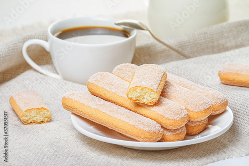 Traditional savoiardi biscuits or ladyfingers cookies on a white plate and a white cup of coffee with a spoon on a napkin. Selective focus