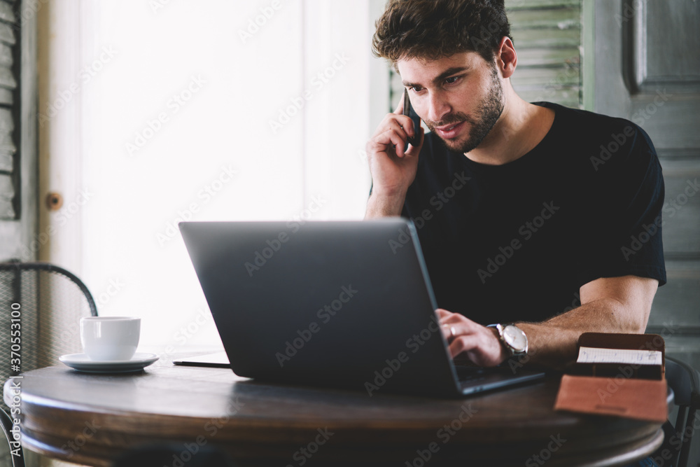 Bearded hipster guy sitting at modern laptop computer connecting to wireless internet while talking on digital cellular during leisure time in cafeteria.Student communicating on cellphone indoors Bearded hipster guy sitting at modern laptop computer connecting to wireless internet while talking on digital cellular during leisure time in cafeteria.Student communicating on cellphone indoors