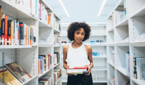 Portrait of attractive dark skinned woman student spending free time in library holding favorite fiction books, beautiful curly african american female looking at camera standing near shelves in store photo