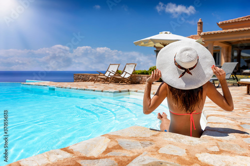 Eine attraktive Frau im Bikini und mit weißem Hut sitzt am Rande des Pooles bei Ihrer Luxus Villa und genießt den Sommertag