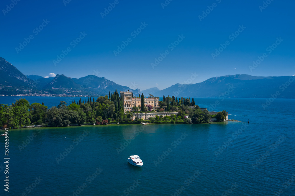 White yacht on the water. The Venetian neo gothic Villa Borghese Cavazza. Aerial view of the island Garda, Lake Garda, Italy. Is the biggest island on Lake Garda. In the background Alps, blue sky.