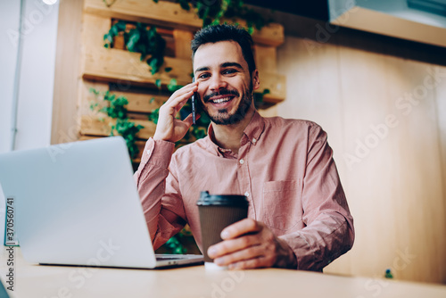 Portrait of cheerful male student dressed in casual look using 4g wireless internet for making positive smartphone conversation with best friend via mobile application while enjoying coffee time