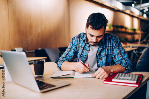 Serious young male student writing essay in textbook for education while sitting at wooden desktop with modern laptop computer and spending time for knowledge and autodidact in university campus