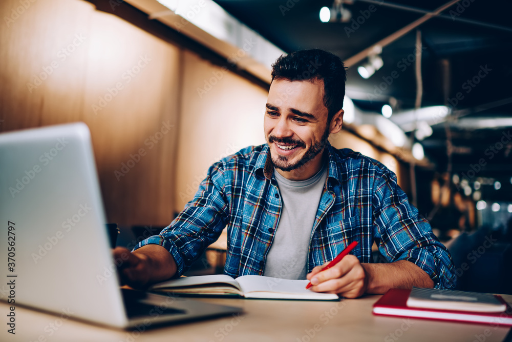 Cheerful hipster guy watching funny movie online and laughing from jokes while sitting in university campus and spending time for e learning and making research of information for course work Cheerful hipster guy watching funny movie online and laughing from jokes while sitting in university campus and spending time for e learning and making research of information for course work