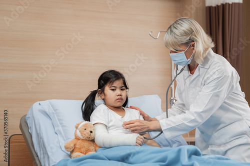Doctor pediatrician examining a little girl patient on bed with teddy bear by stethoscope