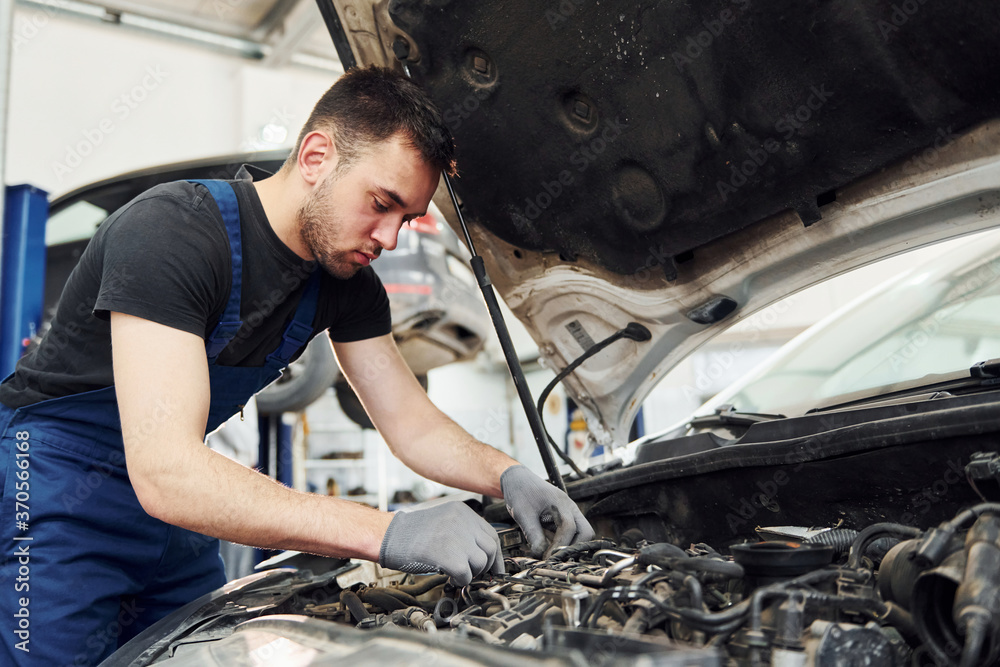 Under the hood. Man in work uniform repairs white automobile indoors. Conception of automobile service Under the hood. Man in work uniform repairs white automobile indoors. Conception of automobile service