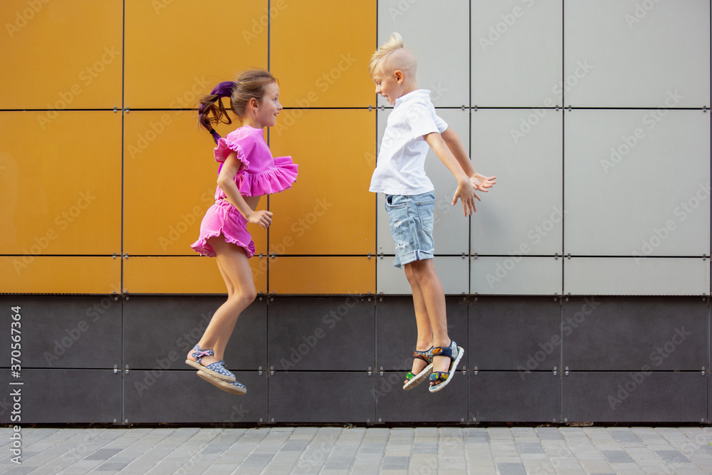 Summertime. Two smiling kids, boy and girl running together in town, city in summer day. Concept of childhood, happiness, sincere emotions, carefree lifestyle. Little caucasian models in bright