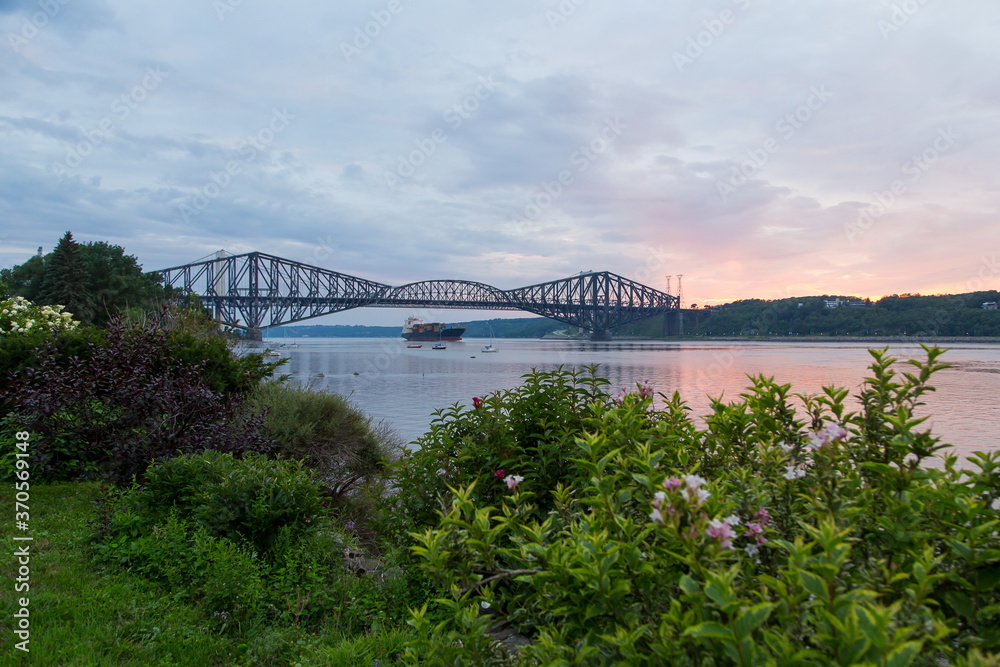 Naklejka premium Sunset view of small boats and large merchant vessel under and near the Quebec Bridge over the St. Lawrence River seen from a south shore garden in soft focus foreground, Lévis, Quebec, Canada