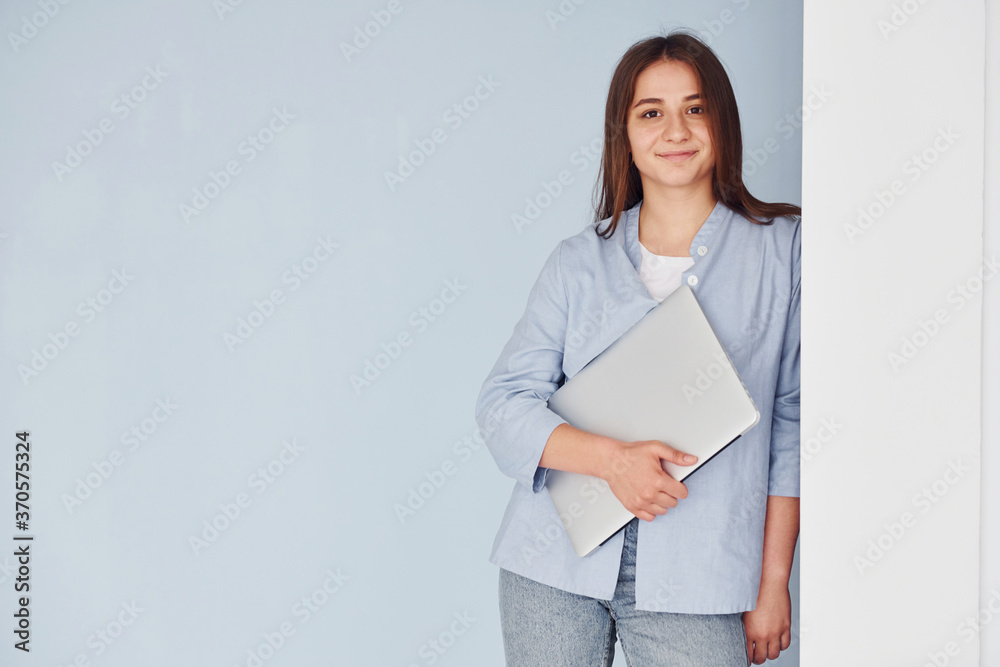 Beautiful young woman in jeans and blue shirt standing against wall with laptop in hands Beautiful young woman in jeans and blue shirt standing against wall with laptop in hands