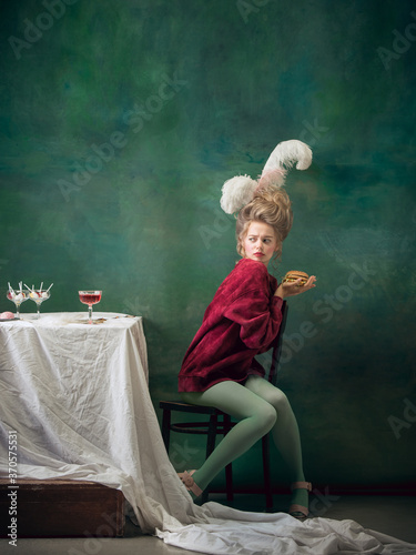 Lovely fast food. Young woman as Marie Antoinette on dark green background. Retro style, comparison of eras concept. Beautiful female model like classic historical character, old-fashioned.