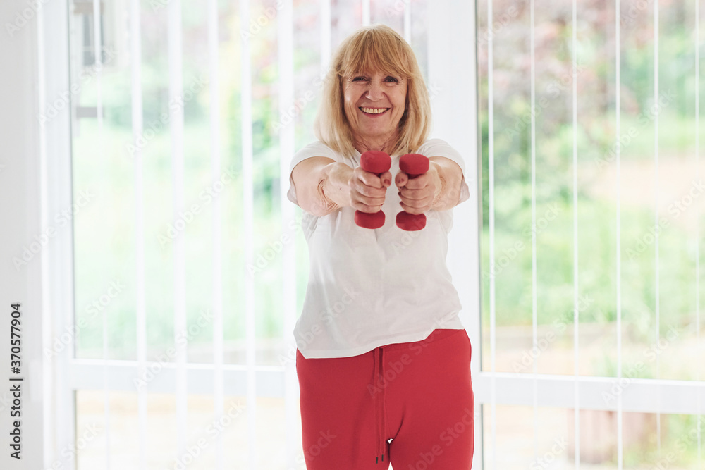 Positive senior woman in sportive clothes indoors at daytime doing exercises with dumbbells Positive senior woman in sportive clothes indoors at daytime doing exercises with dumbbells