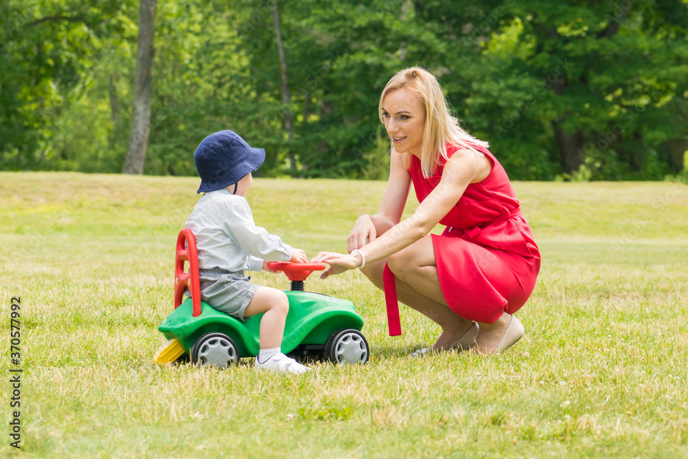 Blonde woman with red dress playing with child on green children car on meadow during summer time