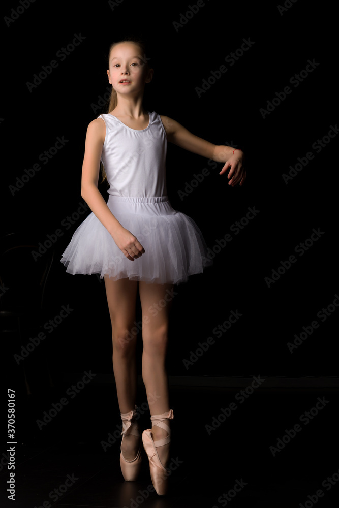 Naklejka premium Cute little girl in a tutu and pointe shoes is dancing in the studio on a black background.
