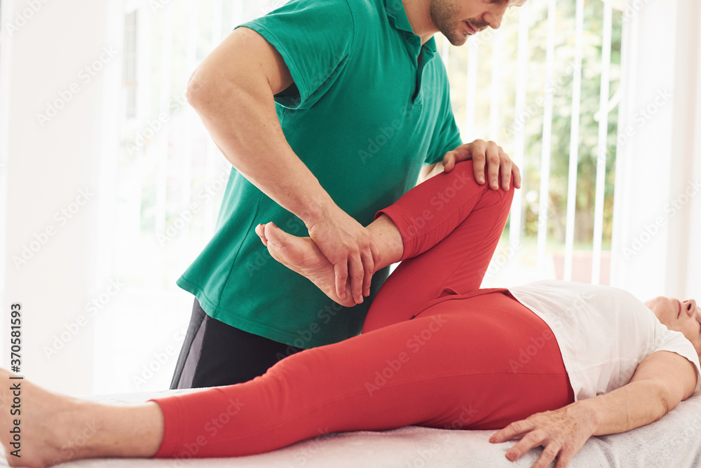 Older woman have a visit in clinic, lying down and having rehabilitation. Male instructor helping her Older woman have a visit in clinic, lying down and having rehabilitation. Male instructor helping her