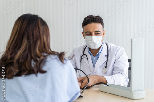 Male doctor wearing face mask using blood pressure gauge with hand woman patient while sitting on the office at the hospital.