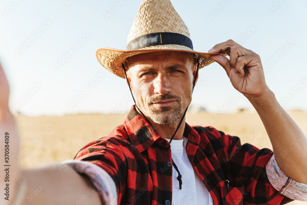 Fototapeta premium Image of adult man taking selfie photo while standing at cereal field