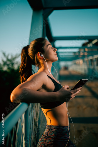 Young sporty woman relaxing and listening music after workout on sunset