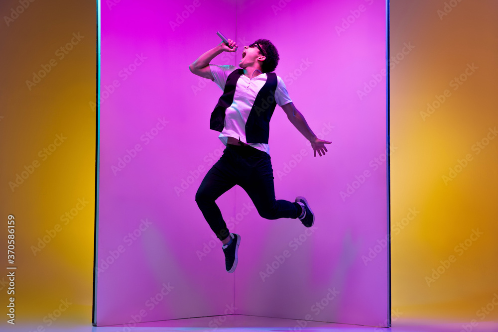 In high jump. Young male musician, singer performing on pink-orange background in neon light. Concept of music, hobby, festival, entertainment, emotions. Joyful party host, singer, portrait of artist.