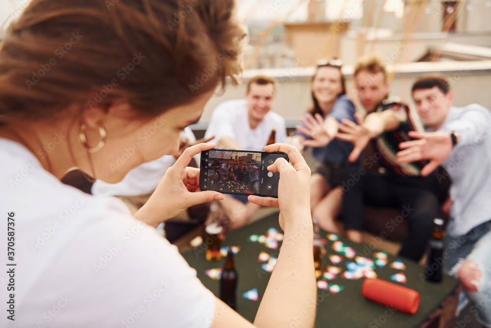 Girl doing photo when people playing card game. Group of young people in casual clothes have a party at rooftop together at daytime
