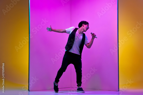 Celebrity. Young male musician, singer performing on pink-orange background in neon light. Concept of music, hobby, festival, entertainment, emotions. Joyful party host, singer, portrait of artist.