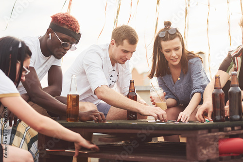 Playing game and sitting by the table. Group of young people in casual clothes have a party at rooftop together at daytime