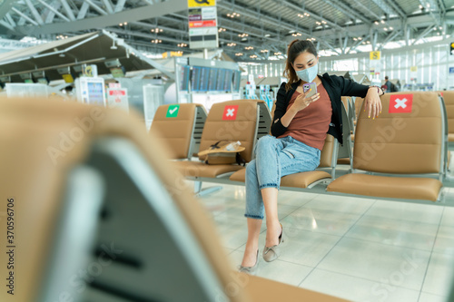 New normal and social distance concept.Asian young woman tourist wearing mask using smartphone searching airline flight status and sitting with distance during corona virus 2019 outbreak at airport