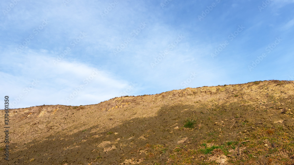 The slope of a clay hill against the backdrop of a beautiful blue sky. Lifeless soil. Sunny weather. Landscape.