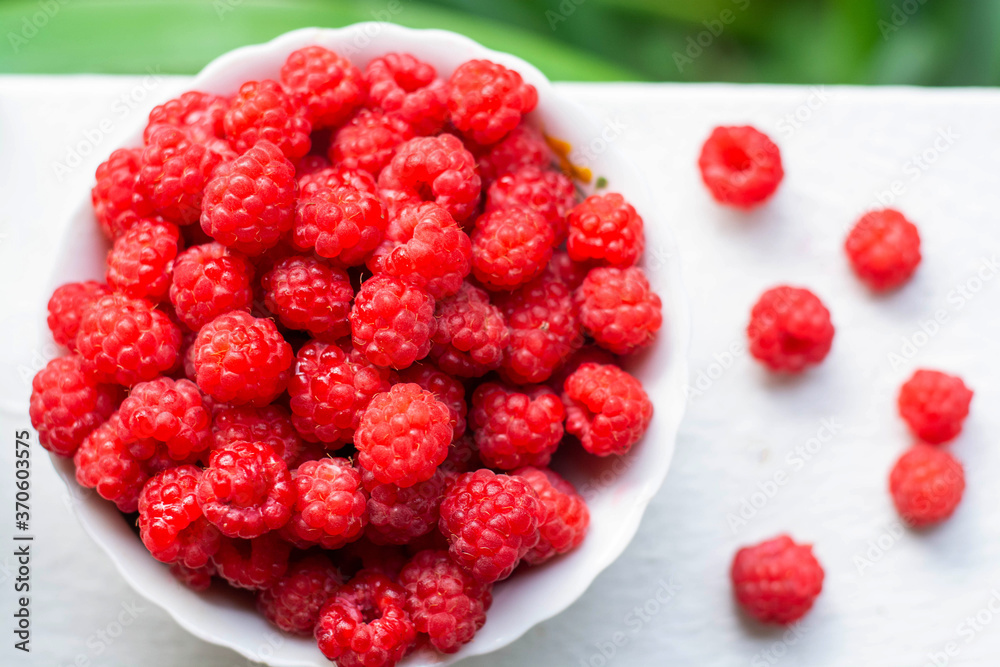 A white cup with fresh ripe raspberries stands against a background of bushes. Natural organic. A white cup with fresh ripe raspberries stands against a background of bushes. Natural organic.