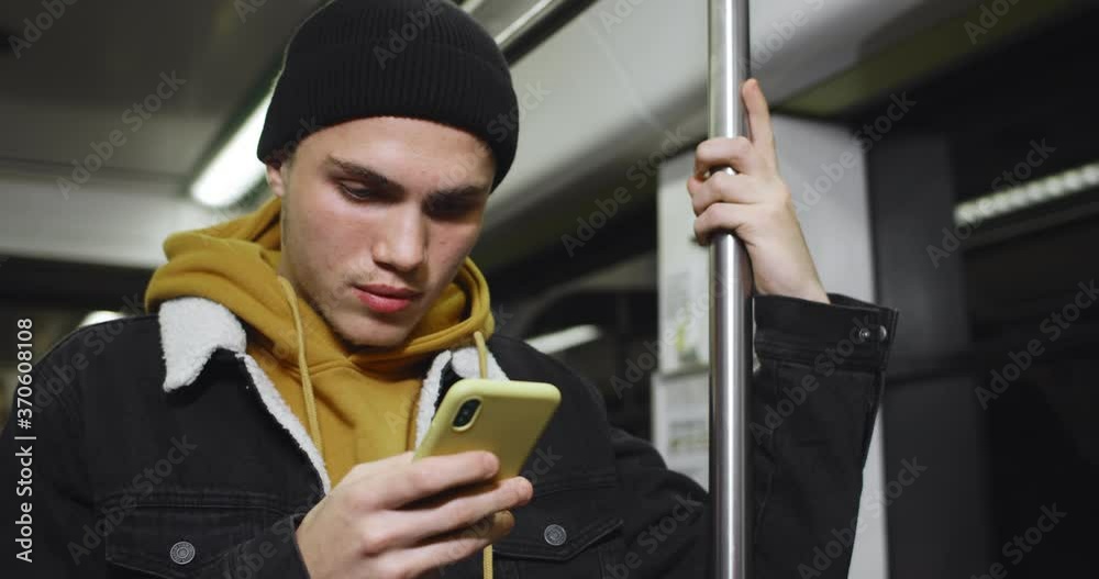 Young handsome student using smartphone and smiling while commuting on public transport. Millennial guy looking at phone screen while typing message in social media.