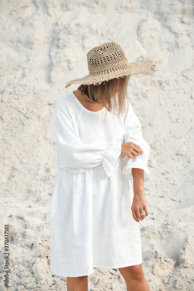 Stylish girl in trendy summer linen dress straw hat posing on the sand. Boho style