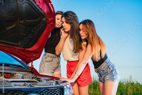 Three confused sexy female friends stuck at road, check red vehicle hood during trip at highway © o_lypa