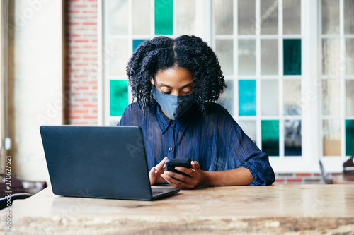Black woman working at cafe on laptop and phone with face mask during covid-19 photo