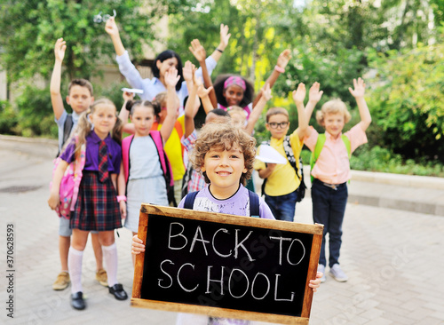 Obraz na plátně a small curly-haired child schoolboy smiles and holds a wooden sign with the inscription Back to school against the background of a group of children of the same class and a female teacher
