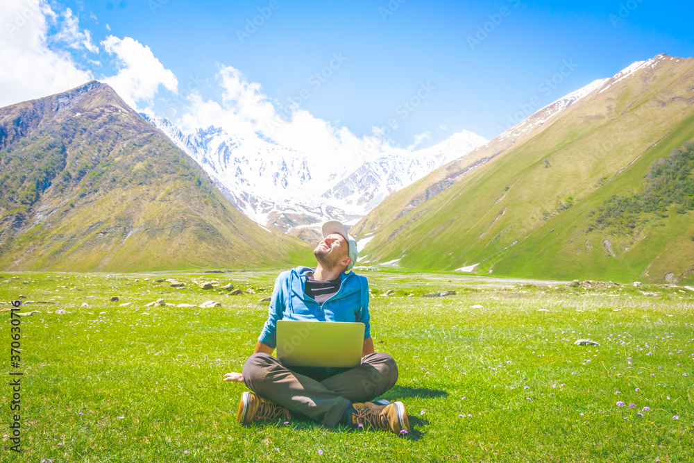 Middle age male person sits with crossed legs on the grass and enjoys the sunsine with grey laptop on his knee. Remote work and self isolation in nature.