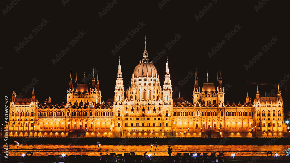Panoramic background orange  night image of Budapest parlament from the  front side with boat and tourist taking a photo of the city. Travel in hungary and historical sites.