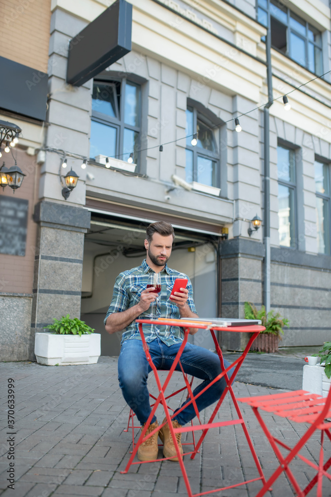 Bearded handsome male drinking red wine, checking his phone in open air cafe