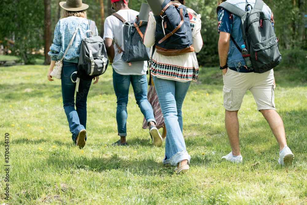 Rear view of young tourists walking on grass and carrying satchels while hiking together Rear view of young tourists walking on grass and carrying satchels while hiking together