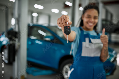 It is done. Young african american woman, professional female mechanic smiling at camera, giving car key and showing thumbs up in auto repair shop