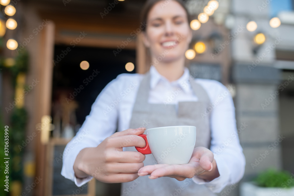 Cute waitress in grey apron offering cup of coffee Cute waitress in grey apron offering cup of coffee