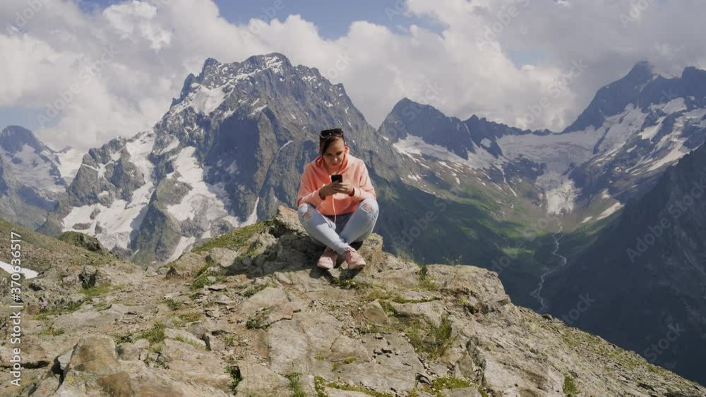 Female traveler using smartphone in mountains. Full body woman sitting on rock and browsing smartphone against cloudy sky on sunny day in mountains Female traveler using smartphone in mountains. Full body woman sitting on rock and browsing smartphone against cloudy sky on sunny day in mountains